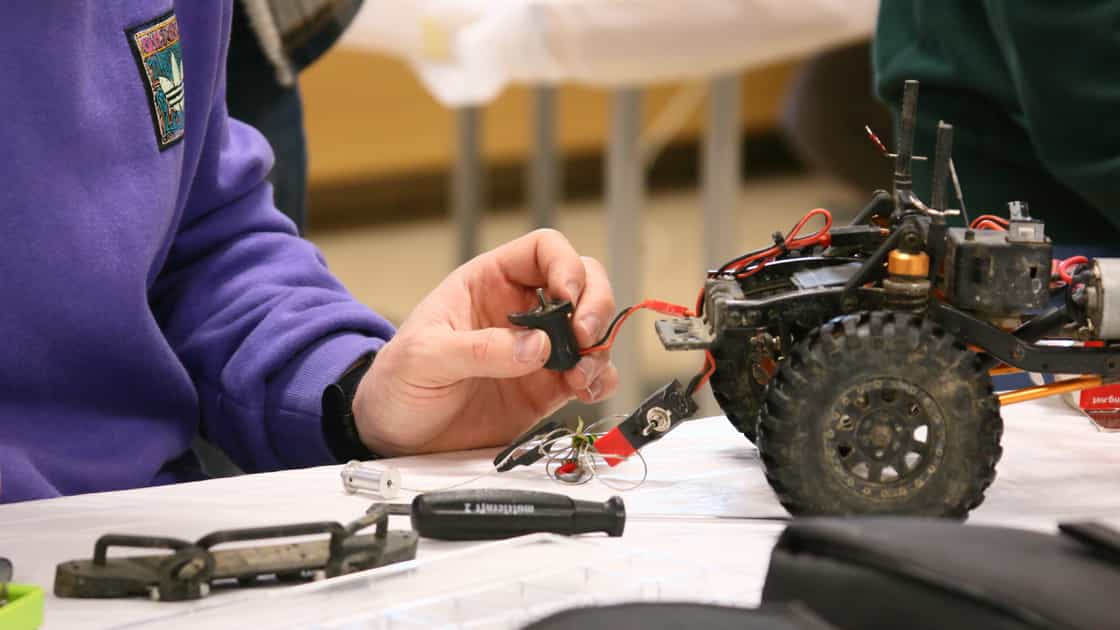 A male person with a lavender-coloured jumper holds the engine of an RC car in his hand and tries to repair the car.