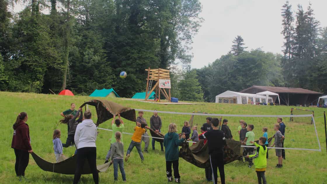 Kinder und Jugendliche spielen Volleyball mit einer Blache auf einem Lagerplatz.